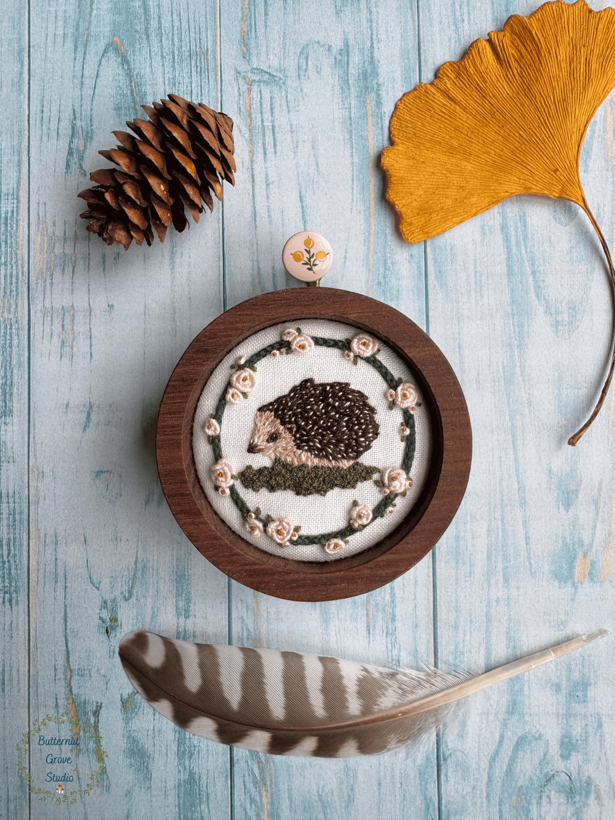 Nestled inside a handmade walnut frame, this miniature artwork features an adorable little hedgie wandering among the moss and blooming roses.

Perfect for a cozy little nook, gallery wall, bookshelf, or anywhere that could use a sprinkling of woodland wonder.

Dimensions: 2" in diameter Hand-stitched embroidery, hand-carved walnut frame

Please note: This piece includes a custom, hand-painted picture hanger, specially finished to complement the artwork (pink background with yellow berries).
