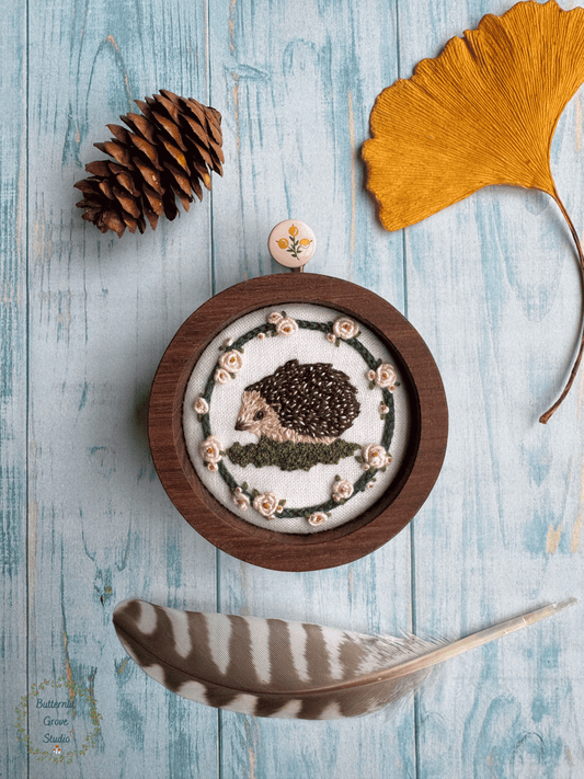 Nestled inside a handmade walnut frame, this miniature artwork features an adorable little hedgie wandering among the moss and blooming roses.

Perfect for a cozy little nook, gallery wall, bookshelf, or anywhere that could use a sprinkling of woodland wonder.

Dimensions: 2" in diameter Hand-stitched embroidery, hand-carved walnut frame

Please note: This piece includes a custom, hand-painted picture hanger, specially finished to complement the artwork (pink background with yellow berries).