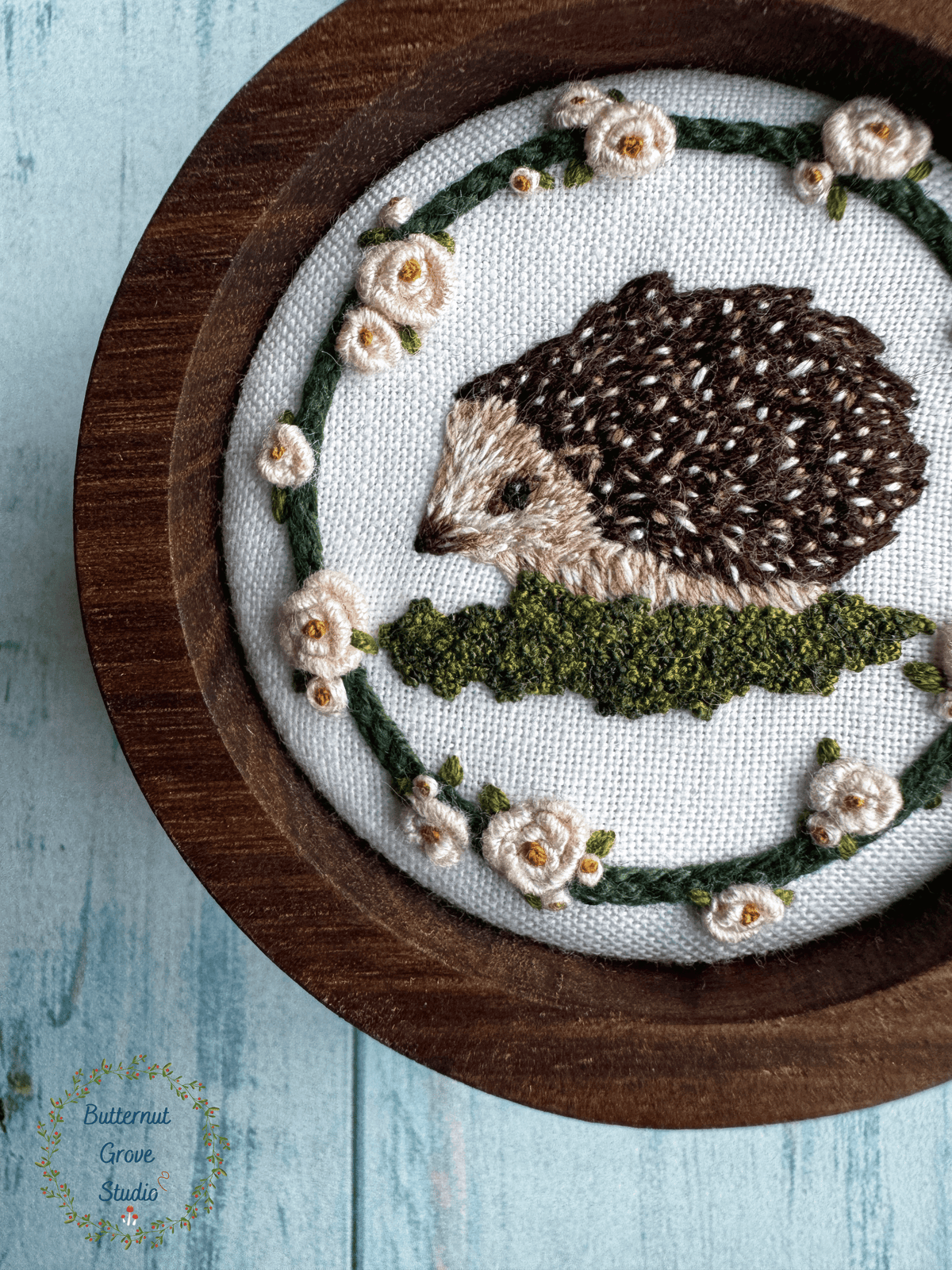 Nestled inside a handmade walnut frame, this miniature artwork features an adorable little hedgie wandering among the moss and blooming roses.

Perfect for a cozy little nook, gallery wall, bookshelf, or anywhere that could use a sprinkling of woodland wonder.

Dimensions: 2" in diameter Hand-stitched embroidery, hand-carved walnut frame

Please note: This piece includes a custom, hand-painted picture hanger, specially finished to complement the artwork (pink background with yellow berries).