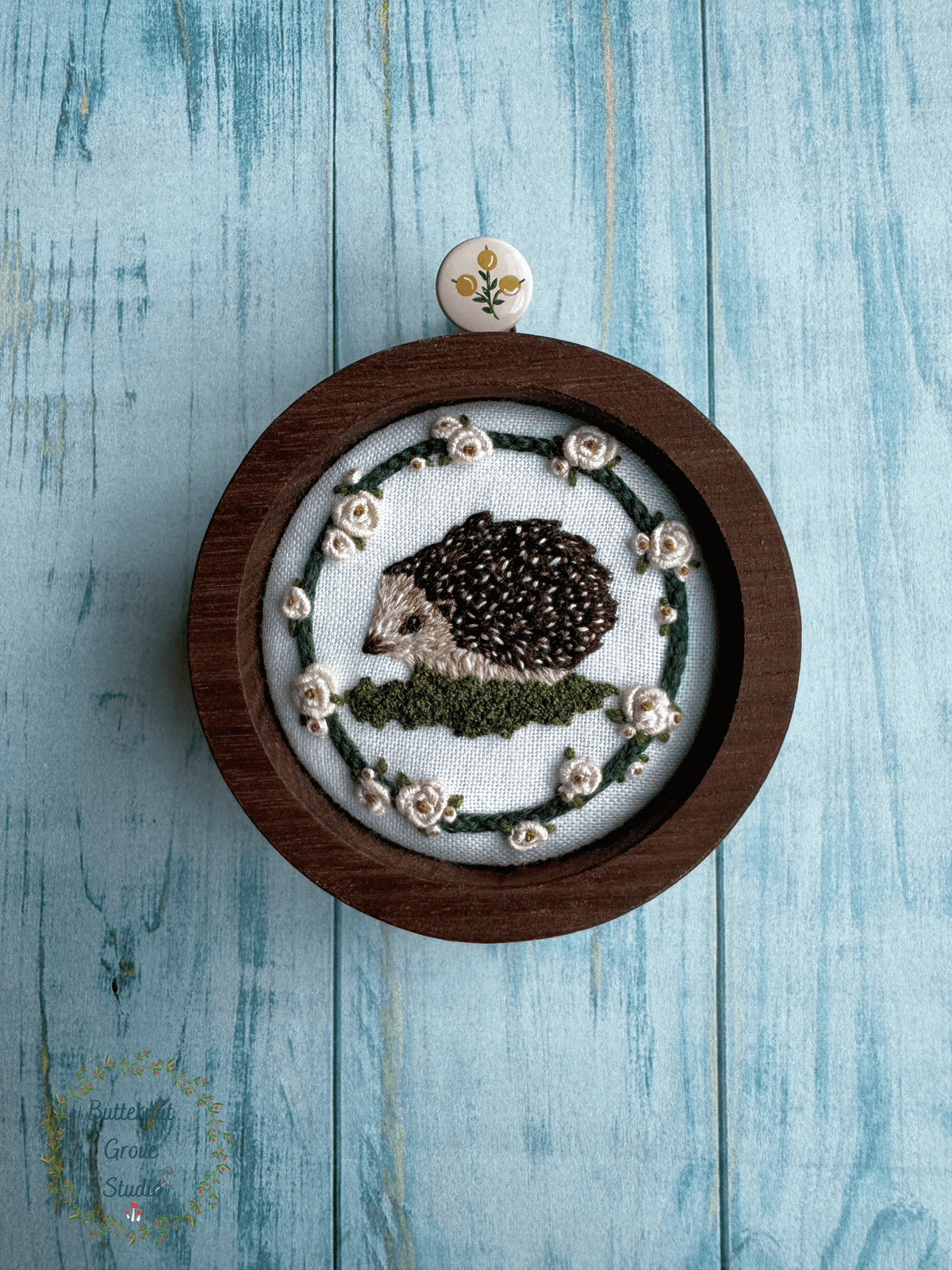 Nestled inside a handmade walnut frame, this miniature artwork features an adorable little hedgie wandering among the moss and blooming roses.

Perfect for a cozy little nook, gallery wall, bookshelf, or anywhere that could use a sprinkling of woodland wonder.

Dimensions: 2" in diameter Hand-stitched embroidery, hand-carved walnut frame

Please note: This piece includes a custom, hand-painted picture hanger, specially finished to complement the artwork (pink background with yellow berries).