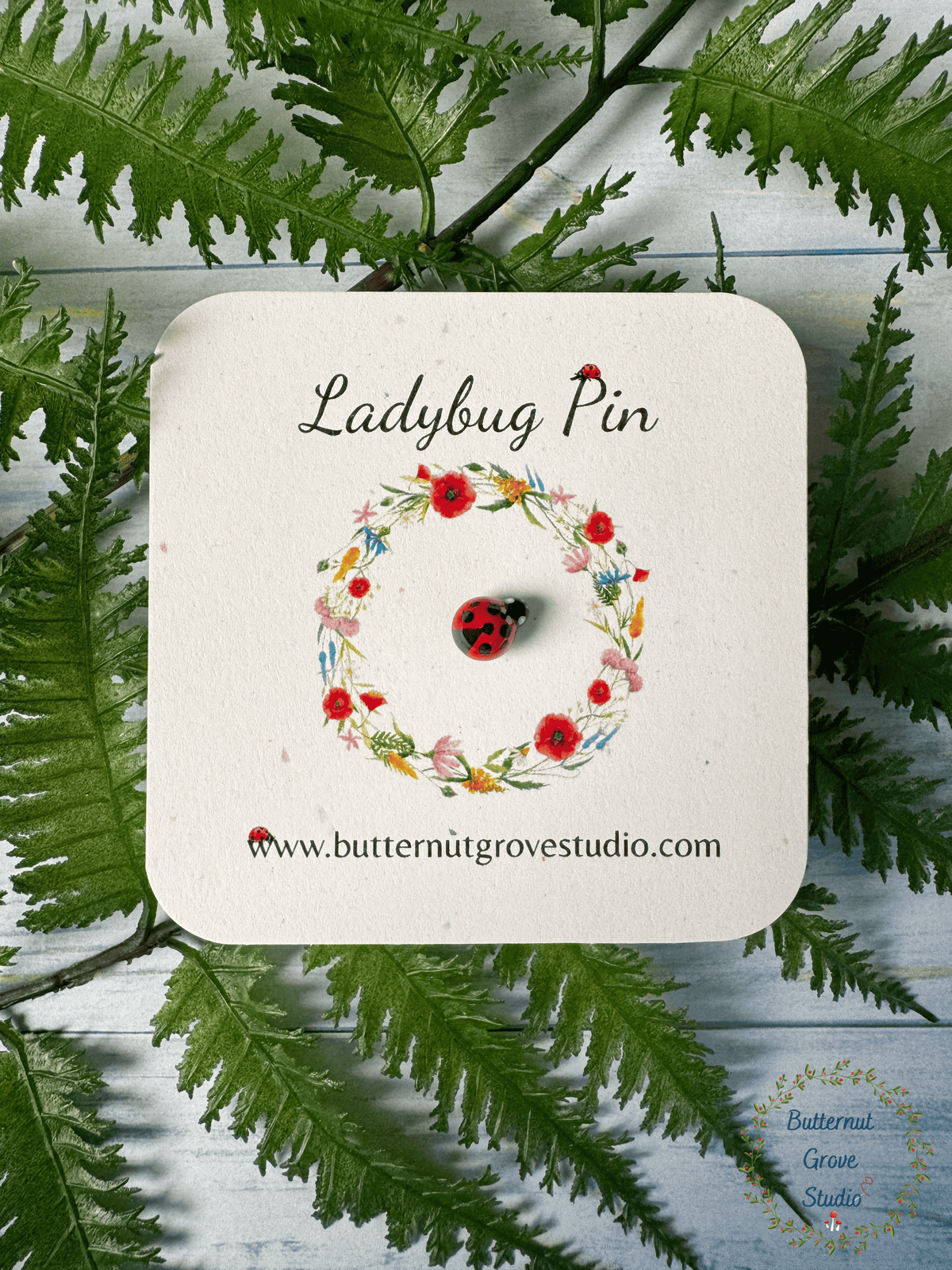 A ladybug pin featuring a small red ladybug with black spots. Ladybug is on a small card, surrounded by a printed floral wreath with the words 'Ladybug Pin' at the top. 