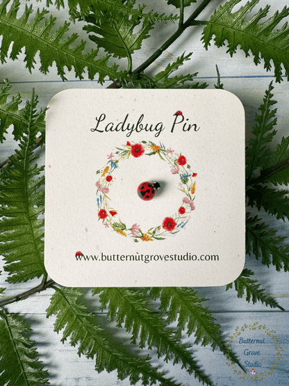 A ladybug pin featuring a small red ladybug with black spots. Ladybug is on a small card, surrounded by a printed floral wreath with the words 'Ladybug Pin' at the top. 