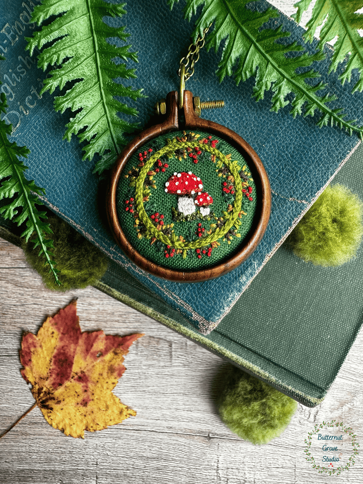 A tiny  1.5" in diameter woodland scene, stitched by hand in delicate detail.  Two bright fly agaric mushrooms sit among a delicate wreath of leaves and berries, all nestled against deep forest-green fabric. Framed in a warm, handmade walnut frame, this little piece has a quiet, fairytale charm, perfect as a miniature work of art for your wall, or a special keepsake wear.
