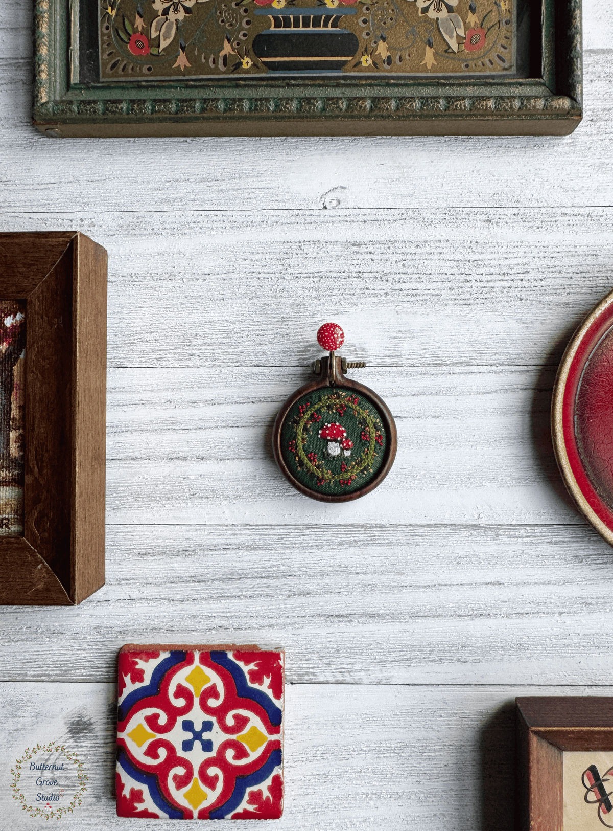 A tiny  1.5" in diameter woodland scene, stitched by hand in delicate detail.  Two bright fly agaric mushrooms sit among a delicate wreath of leaves and berries, all nestled against deep forest-green fabric. Framed in a warm, handmade walnut frame, this little piece has a quiet, fairytale charm, perfect as a miniature work of art for your wall, or a special keepsake wear.