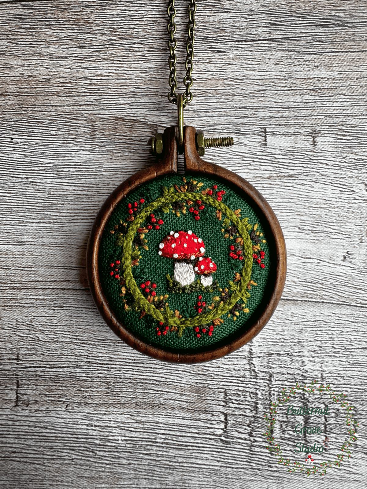 A tiny  1.5" in diameter woodland scene, stitched by hand in delicate detail.  Two bright fly agaric mushrooms sit among a delicate wreath of leaves and berries, all nestled against deep forest-green fabric. Framed in a warm, handmade walnut frame, this little piece has a quiet, fairytale charm, perfect as a miniature work of art for your wall, or a special keepsake wear.