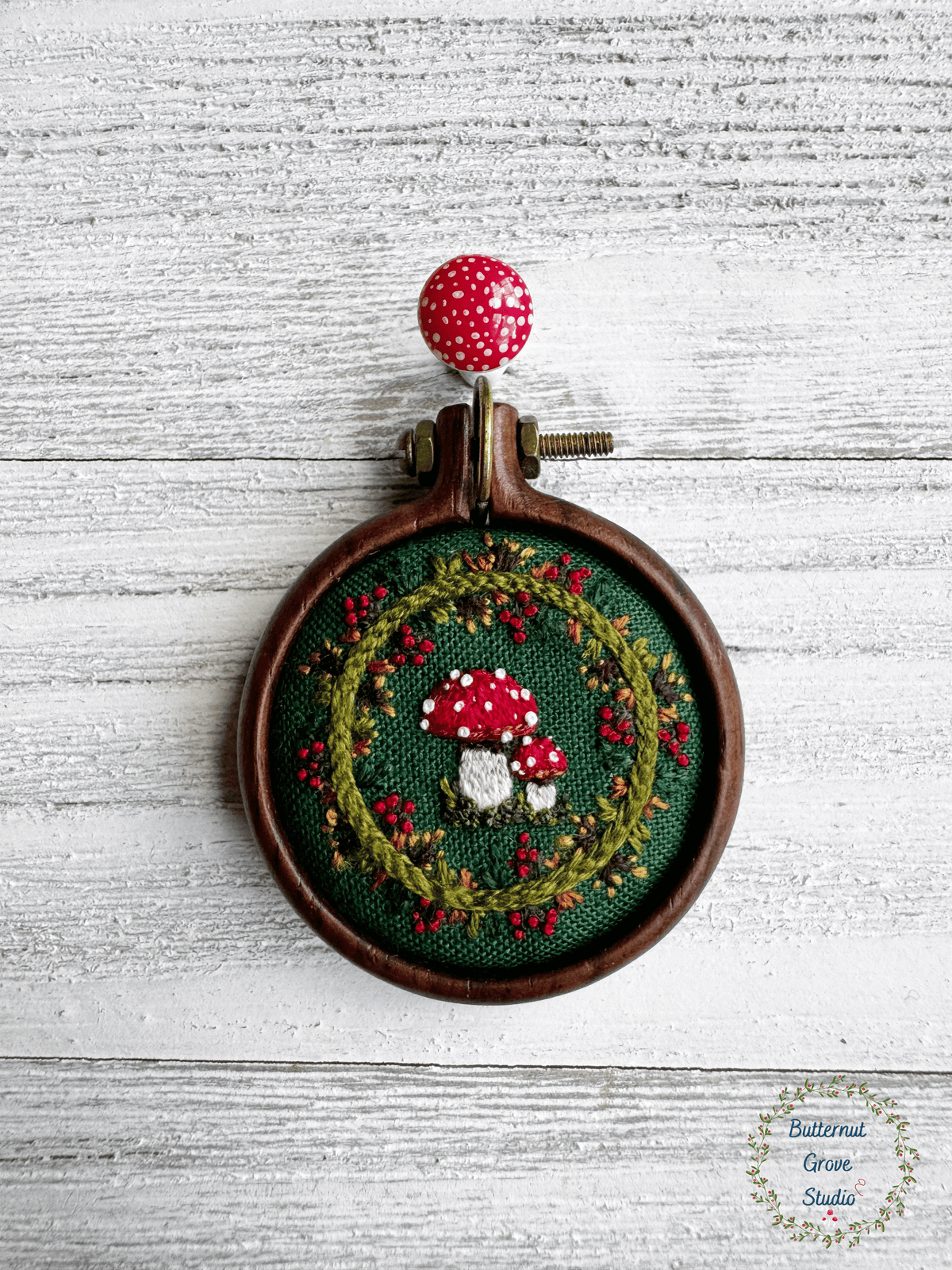 A tiny  1.5" in diameter woodland scene, stitched by hand in delicate detail.  Two bright fly agaric mushrooms sit among a delicate wreath of leaves and berries, all nestled against deep forest-green fabric. Framed in a warm, handmade walnut frame, this little piece has a quiet, fairytale charm, perfect as a miniature work of art for your wall, or a special keepsake wear.