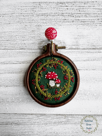 A tiny  1.5" in diameter woodland scene, stitched by hand in delicate detail.  Two bright fly agaric mushrooms sit among a delicate wreath of leaves and berries, all nestled against deep forest-green fabric. Framed in a warm, handmade walnut frame, this little piece has a quiet, fairytale charm, perfect as a miniature work of art for your wall, or a special keepsake wear.