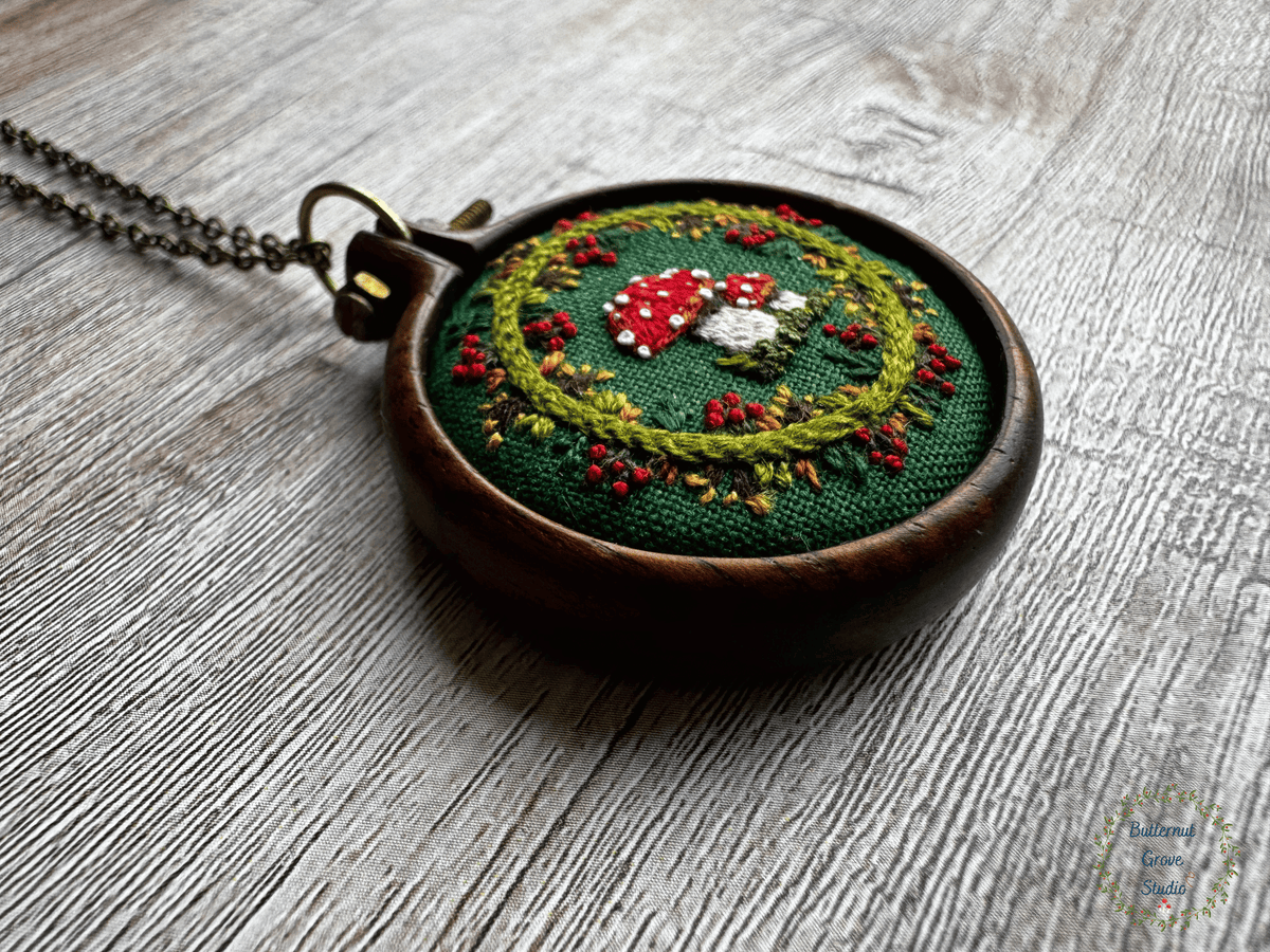 A tiny  1.5" in diameter woodland scene, stitched by hand in delicate detail.  Two bright fly agaric mushrooms sit among a delicate wreath of leaves and berries, all nestled against deep forest-green fabric. Framed in a warm, handmade walnut frame, this little piece has a quiet, fairytale charm, perfect as a miniature work of art for your wall, or a special keepsake wear.