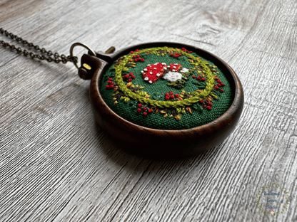 A tiny  1.5" in diameter woodland scene, stitched by hand in delicate detail.  Two bright fly agaric mushrooms sit among a delicate wreath of leaves and berries, all nestled against deep forest-green fabric. Framed in a warm, handmade walnut frame, this little piece has a quiet, fairytale charm, perfect as a miniature work of art for your wall, or a special keepsake wear.